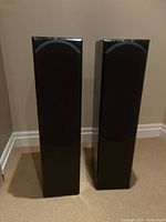 Pair of Kinima black floor-standing speakers with black fabric grill covers in place, standing on carpet.