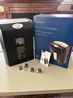 Black Insignia micro-cut shredder, original box, manual, and four caster wheels sealed in plastic bags shown on table.