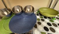 Photo showing three stainless steel snack bowls placed on green and blue woven placemats next to a green metal serving bowl and square polka-dotted placemats.