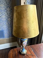 Porcelain vase lamp with gold tapered lampshade on a wooden surface against wallpapered wall.