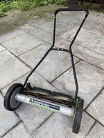 Full front angle view of American reel type lawn mower on stone patio, showing handlebar frame, wheels, and cutting reel.