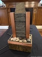 Front view of the water fountain showing the textured stone centerpiece framed by copper-colored sides with natural stones at base.