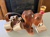 Side angle showing brown ceramic dog decanter holding six small ceramic cups on pegs along its body, cork stopper on nose.