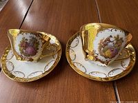 Photo showing pair of demitasse cups and saucers with detailed gold decoration and romantic couple motifs.