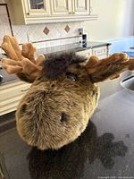 Close-up image of the whimsical moose head showing dense brown faux fur, plush antlers, tuft on top, and shiny plastic eyes with a black nose.