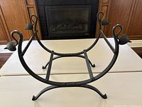 Wrought iron firewood holder displayed on a table in front of a fireplace, showing the full holder structure with curved sides and decorative leaves.