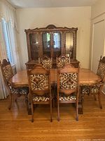 Photo showing the ornate solid oak dining table with six carved wood and upholstered chairs arranged around it, hardwood flooring visible.