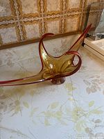 Side view showing the sculptural form, amber color, and red edge trim of the glass vase on a patterned surface with vintage wallpaper in background.