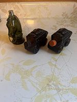 Photo of two brown glass carriage-shaped bottles with orange screw caps and one brown glass horse head bottle with silver screw cap and metal bridle detail on a floral patterned surface.