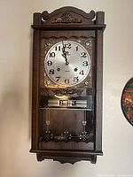 Front view of the Badrad vintage wood and metal wall clock showing dial, decorative carvings, and day-date display