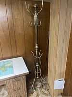 Full view of the tall brass coat rack placed next to a desk and wooden paneled wall.