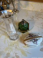 Photo showing clear glass vase, green hexagonal glass lighter, and oval floral ceramic trinket box on a patterned surface.