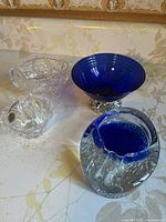 Photo showing four glass bowls/dishes in blue and clear colors on a floral patterned surface, highlighting the different designs and materials.