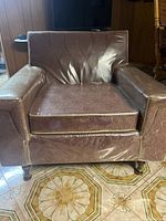 Front view of brown fabric armchair covered with protective plastic showing paisley upholstery, padded arms and seat cushion, and turned wood legs.