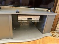 Front view of gray TV stand showing top surface with black remote, two glass shelves holding a stereo component, composite frame on sides and base.