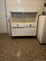 Front view of the kitchen storage cabinet showing sliding glass doors on the top compartment, two drawers with round handles, and lower covered sliding doors. Visible wear and discoloration inside the glass compartment and on cabinet surfaces.