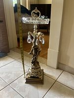 Full view of the ornate decorative stand showing height with measuring tape, brass base with sculpted cherub figure, and crystal plate top with handle.