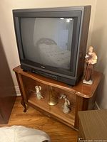 RCA CRT TV on wooden stand with carved edges and glass doors, showing two angel figurines inside and a standing figurine beside TV