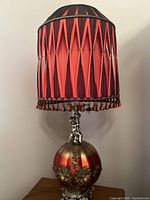 Full image of the Victorian lamp showing the pleated red and black fabric shade with fringe and the spherical base with floral accents and cherub figurine on top.