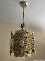 Hanging vintage bronze chandelier with ornate floral embossed panels and faceted crystal clusters inside glass sections, shown suspended by chain from ceiling