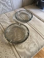 Two clear glass ashtrays placed on a patterned fabric surface. Both have a round shape with three resting grooves on the rim.