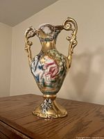 Front perspective of the ceramic vase showing detailed classical-style figures painted on a gold finish with elaborate handle details.