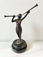 Front and left side view of bronze statue of a nude lady blowing a trumpet, standing on a round black marble base.