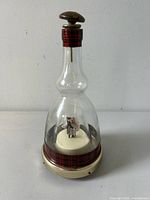 Front view of the clear glass decanter with dancing ballerina couple and plaid band on neck and base, showing its height and top stopper.