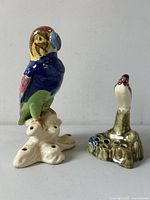 Front and side views of both ceramic bird flower frogs on a gray surface showing their shape, color, and flower stem holes.