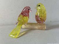 Clear front-right angled view showing two colorful Murano glass love birds perched on a textured cylindrical glass branch, one bird's yellow tail extending downward.