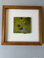 Framed square painting showing greenish background and an abstract object, set within a simple wood frame