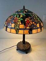 Full view of the Gothic Tiffany style stained glass table lamp illuminated, showing colorful floral and leaf patterned shade and dark metal footed base.