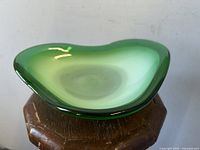 Freeform green hued Coquille art glass bowl displayed on wooden stool with dark to light green gradient and smooth glossy finish.