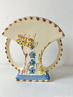 Front view of the cream-colored Art Deco vase decorated with butterfly and blue and yellow flowers, showing the fan-shaped top and handle on right side.