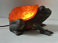 Side view of the figural frog lamp illuminated, showing amber stained glass shell and metal dark body with textured detail.