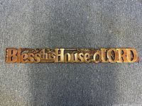 Top-down view of the carved wooden wall sign on a dark surface showing the phrase 'Bless this House o LORD'