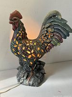 Front view of the ceramic rooster table lamp lit up showing leaf cut-out light pattern