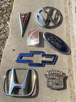 Photo of 7 assorted car emblems including Pontiac, Volkswagen, Saturn, Ford, Chevrolet, Honda, and Cadillac, laid out on a concrete surface.