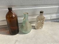 Three vintage glass bottles on a ledge: One brown Javex 32 oz bottle with screw cap, one clear green decorative bottle with handle, and one small clear rectangular bottle with surface residue.