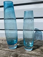 Two blue glass vases placed outdoors on a wooden surface, one taller and one shorter, showing the color and shape.