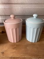 Photo of the two ceramic canisters with lids, pink and blue, side by side on a wooden surface, showing shape and color.