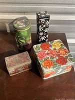 Set of 4 decorative tin boxes on a table: large floral rose square box, tall black snowflake rectangular box, green cat foliage cylindrical box, small vintage red floral rectangular box.
