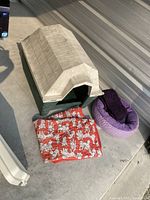 Plastic Ruff Hauz dog house shown on concrete floor next to purple circular dog bed with knitted cover and red dog blanket with white dalmatian prints folded in front.