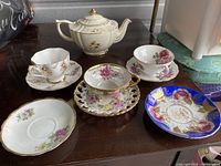 Photo showing white bone china teapot with floral details and five teacups with assorted floral designs on their saucers.