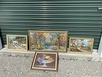 Four framed textile artworks including three cross stitch landscapes and one embroidered fruit bowl, placed on gravel in front of a green door.