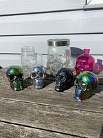 Seven glass skull decorative items displayed on a wooden surface outdoors in natural light. Includes multicolor rainbow finish skulls, clear and black skulls, and a pink skull bottle.