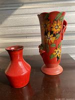 Photo showing two red clay vases, one tall with floral hand-painted design and handle, the other smaller and plain red.