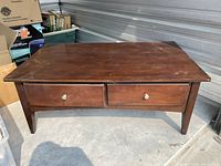 Front view of the wooden coffee table showing two drawers and tapered legs on concrete floor with storage boxes nearby.