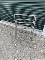 Front view of the modern chrome metal towel rack positioned on gravel ground in front of a green shutter door.