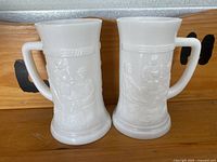 Two white milk glass beer steins side by side, showing embossed figure designs and handles.